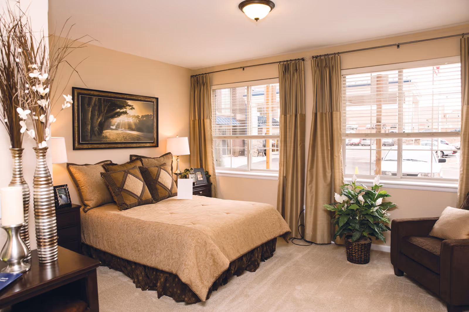Sunny, warmly decorated bedroom with a made bed, bedside tables and lamps, an armchair, and large windows with curtains.