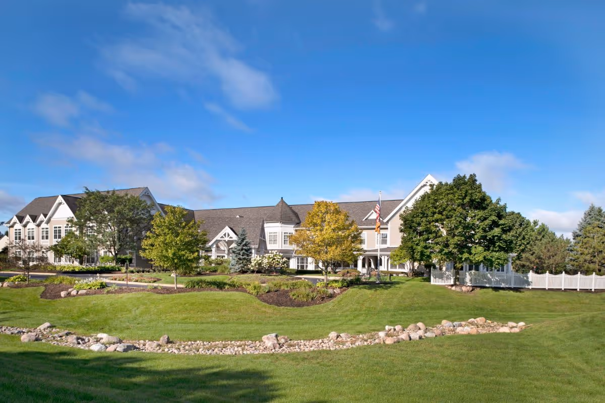 Front exterior of a large senior living facility with landscaped lawns, trees, and an American flag under a blue sky.