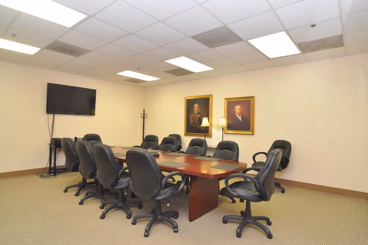 A conference room with a long wooden table surrounded by ten black office chairs on wheels. The room has beige walls with two framed portraits and two standing lamps. A flat-screen TV is mounted on the wall to the left, with cables and a small stand beneath it. The ceiling has recessed lighting panels.
