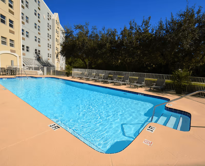 Outdoor swimming pool with clear blue water next to a multi-story building. Several lounge chairs are lined up along the poolside, and there are trees and greenery in the background under a clear blue sky.