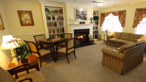 A cozy living room with a lit fireplace, two striped sofas, a wooden table with four chairs, bookshelves filled with books and decorative items, framed artwork on the walls, and windows with floral curtains letting in natural light.