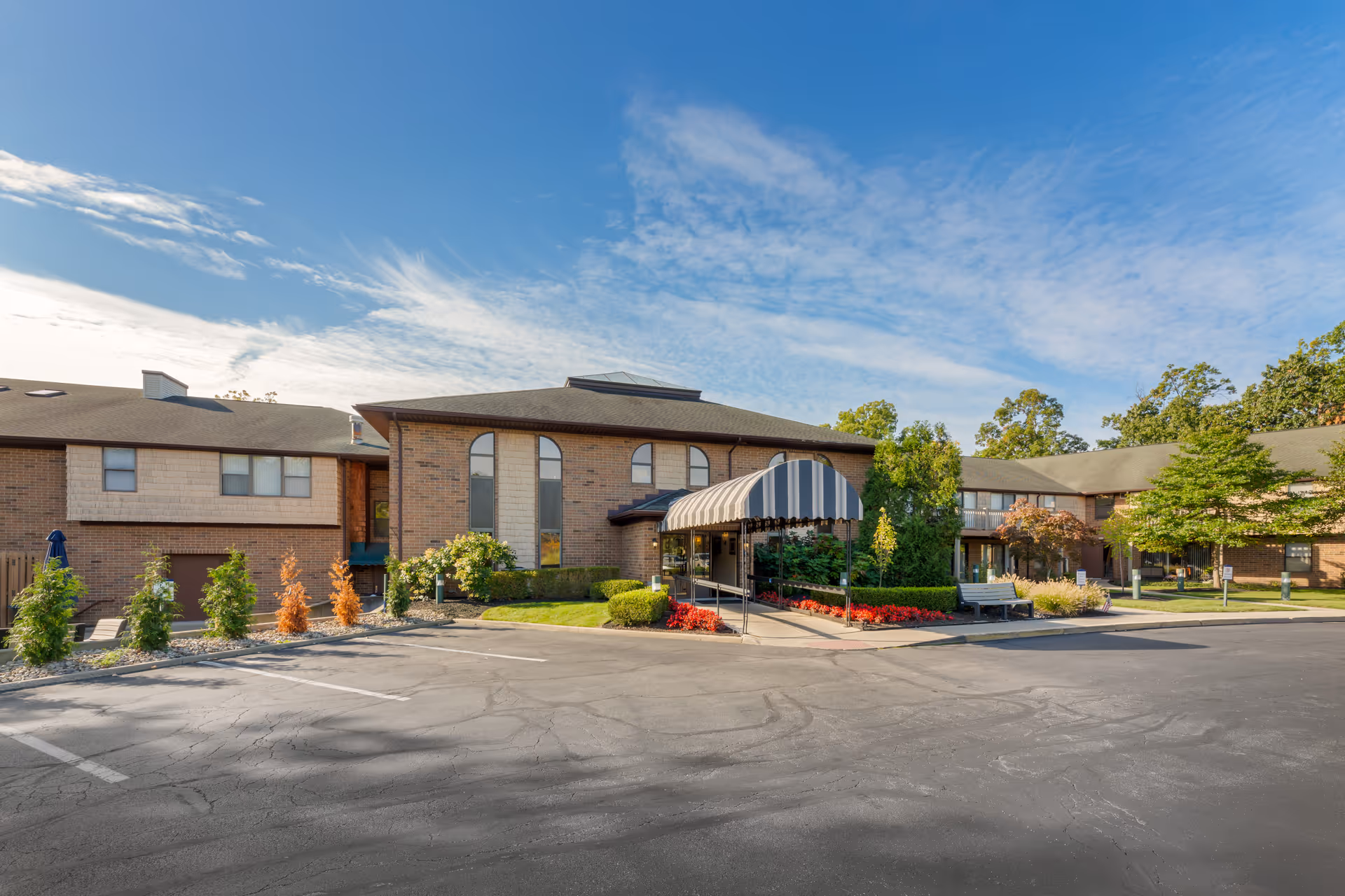 Brick two-story senior living building with a striped entrance canopy, landscaped flower beds and a parking lot under a blue sky.