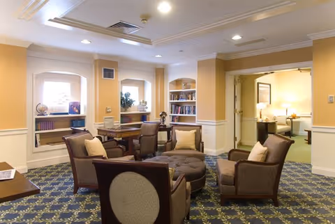 A cozy living room area in a senior living facility with four cushioned armchairs arranged around a large round ottoman. The room has built-in bookshelves with books and decorative items, soft yellow walls with white trim, and patterned carpet flooring. There is a doorway leading to another room with additional seating and warm lighting.