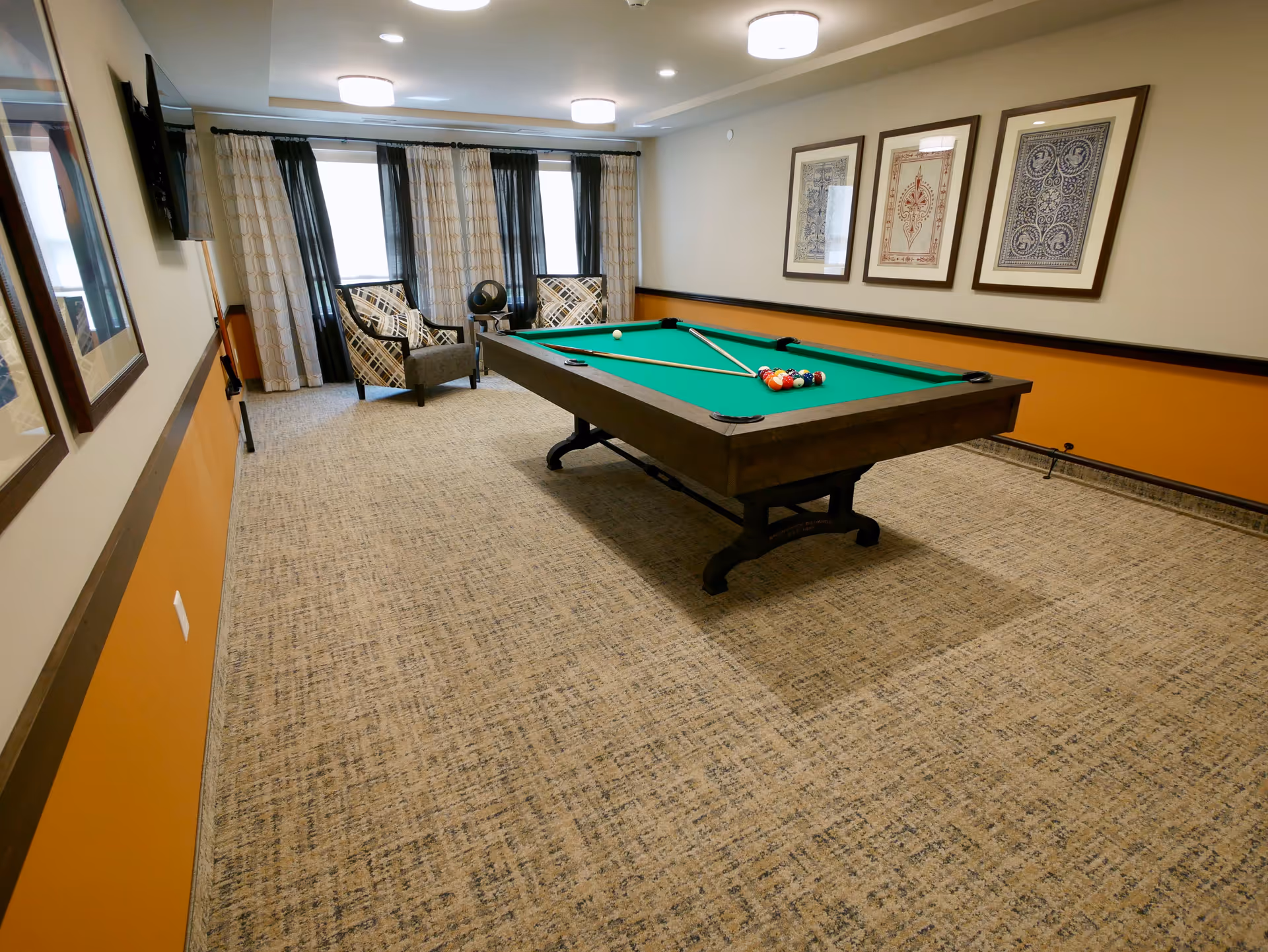 A recreational room with a pool table in the center, two patterned armchairs near the window with curtains, a wall-mounted TV, and three framed artworks on the wall. The room has beige carpet and walls with an orange accent panel.