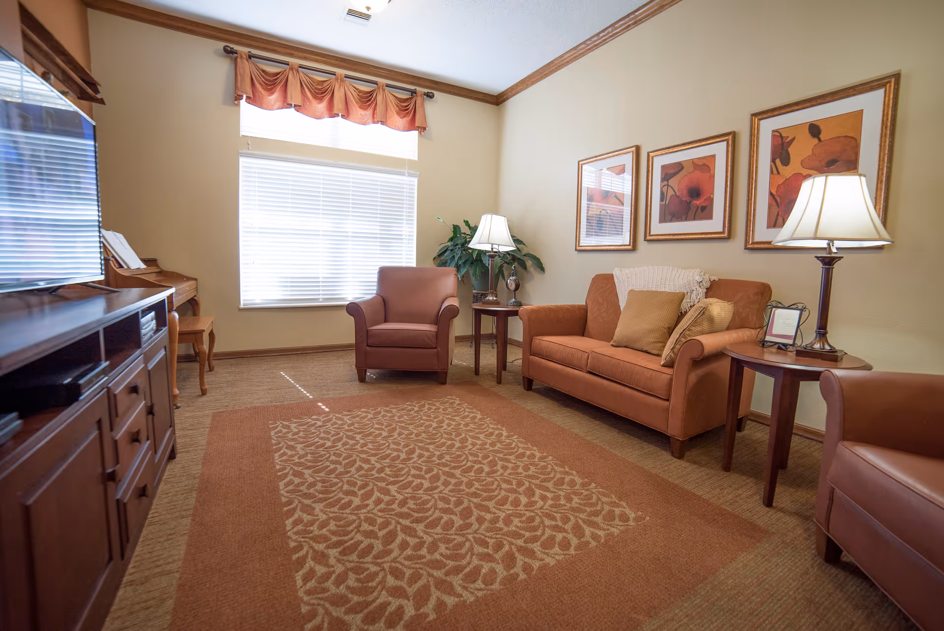 A cozy living room with a brown sofa, two matching armchairs, a patterned area rug, two side tables with lamps, three framed floral artworks on the wall, a large window with blinds and a valance, and a wooden entertainment unit with a TV and a piano in the corner.