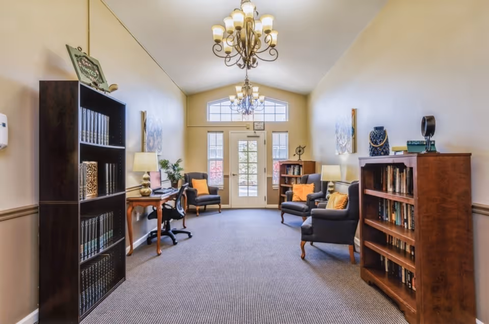 A bright and cozy sitting area with beige walls and carpeted floor. The room features two dark wooden bookshelves filled with books on either side, several comfortable armchairs with orange cushions, a wooden desk with a lamp and computer, and two elegant chandeliers hanging from the ceiling. Large windows and a glass door at the far end allow natural light to fill the space.