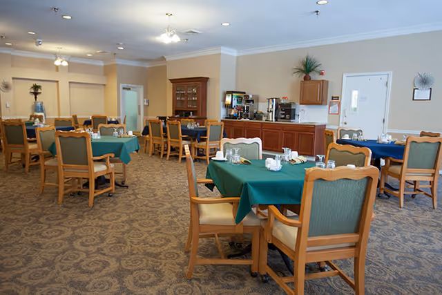 A spacious dining room with multiple wooden tables covered with green and blue tablecloths, each set with glasses, napkins, and condiments. Wooden chairs with green cushions surround the tables. In the background, there is a wooden cabinet and a counter with coffee machines and other beverage equipment. The room has beige walls and patterned carpet flooring.