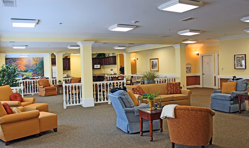 A spacious and well-lit living room area in a senior living facility with multiple comfortable armchairs and sofas arranged around small tables. The room features beige walls, carpeted floors, white railings, and a kitchen area in the background. There are framed pictures on the walls and decorative plants placed around the room.