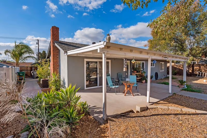 Single-story gray house with a covered patio, outdoor chairs and table, and nearby desert landscaping under a blue sky.