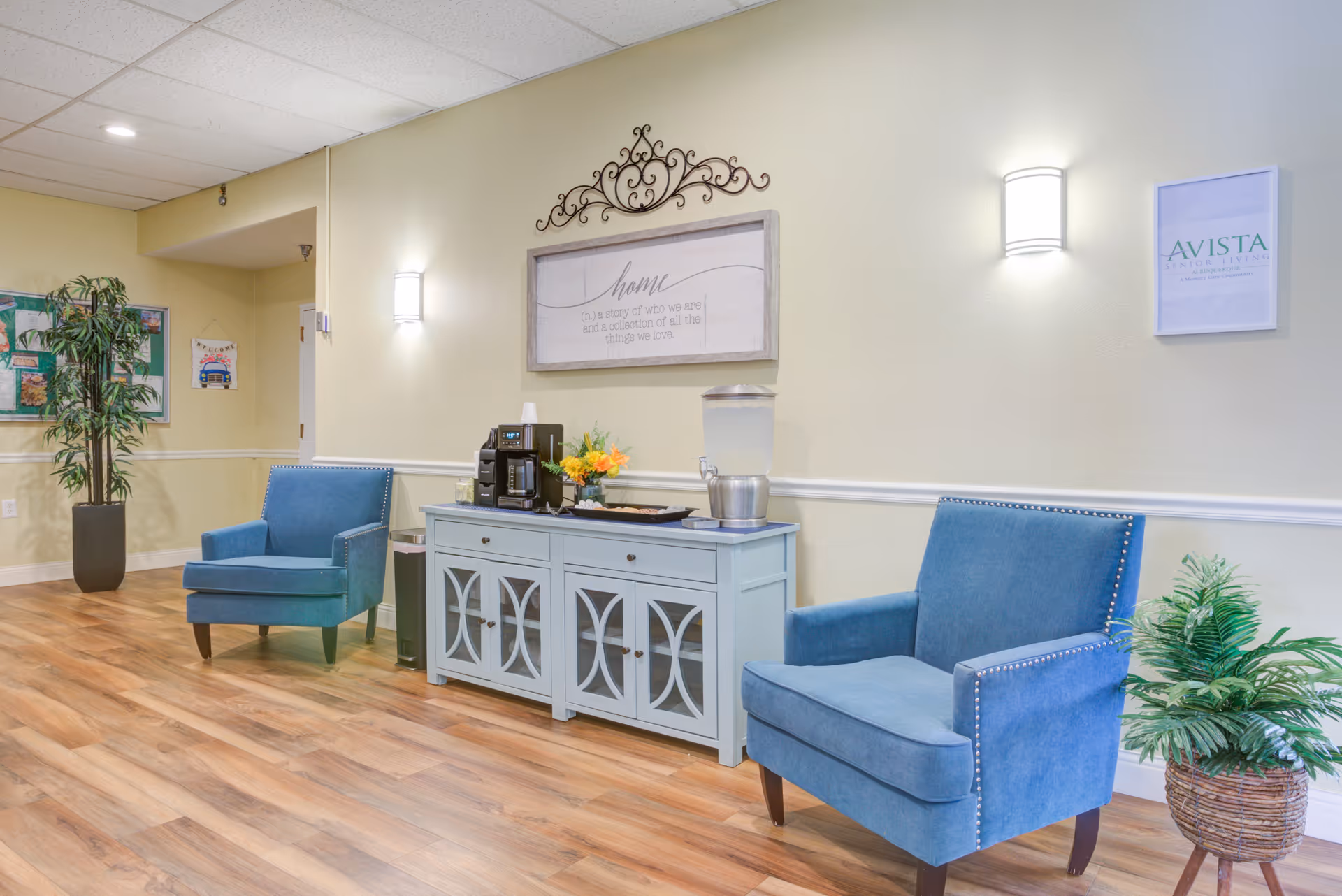 A cozy seating area in a senior living facility with two blue armchairs, a light blue sideboard holding a coffee maker, water dispenser, and a flower arrangement. The walls are light yellow with two wall sconces and decorative wall art, including a framed sign that says 'home' and a framed Avista Senior Living logo. There is a potted plant on the right and a bulletin board with notices on the left.