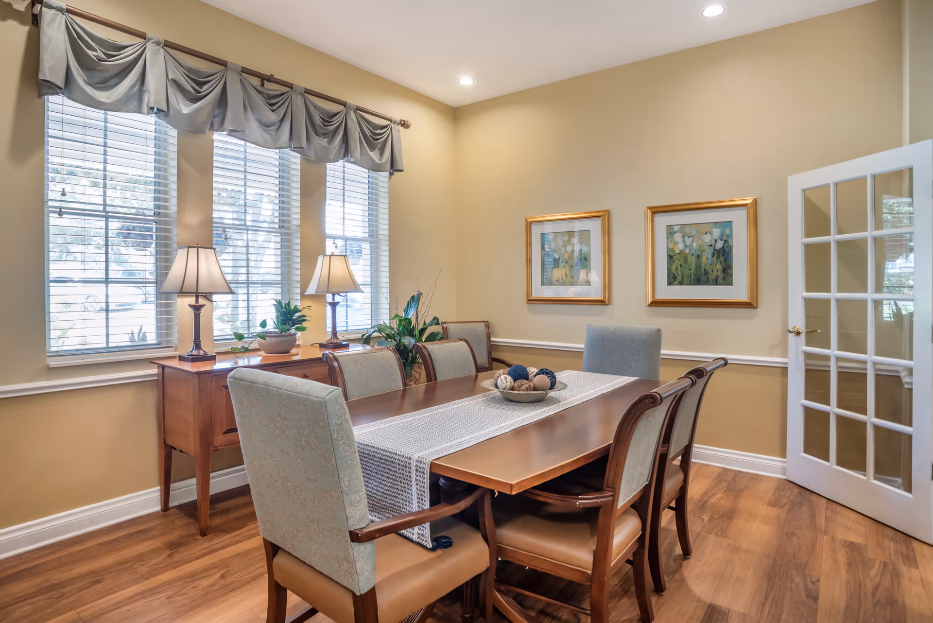 A dining room with a wooden table surrounded by six upholstered chairs. The table has a decorative runner and a bowl with decorative balls. Behind the table, there is a wooden sideboard with two table lamps and a plant. Three windows with blinds and draped valances allow natural light into the room. Two framed floral paintings hang on the beige wall, and a white door with glass panes is partially open.