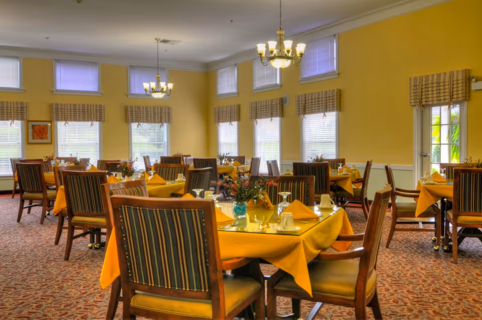 A dining room with multiple tables covered with yellow tablecloths, each set with glasses, cups, and napkins. The chairs have wooden frames with striped upholstery. The room has yellow walls, several windows with blinds and valances, and two chandeliers hanging from the ceiling.