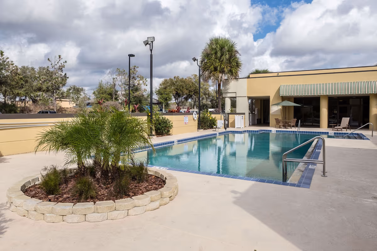 Outdoor swimming pool area at Grand Villa of DeLand with a small circular garden containing palm plants in the foreground. The pool is surrounded by a concrete deck with metal handrails and a building with a striped awning and patio furniture in the background. Trees and cloudy sky are visible beyond the pool area.