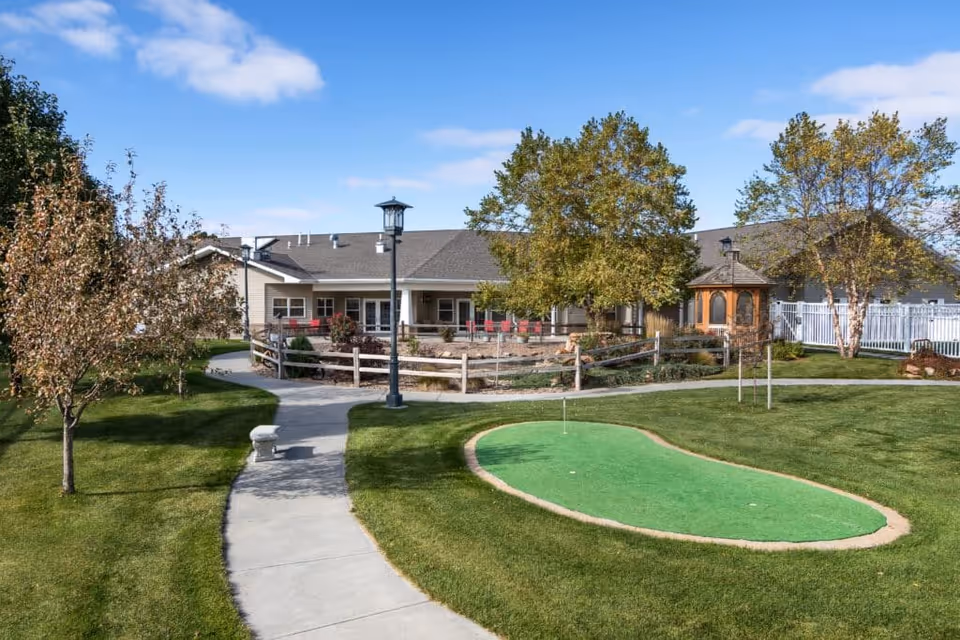 Outdoor view of a senior living facility with a putting green, paved walking path, trees, a gazebo, and a building with a covered porch and red chairs under a blue sky with some clouds.