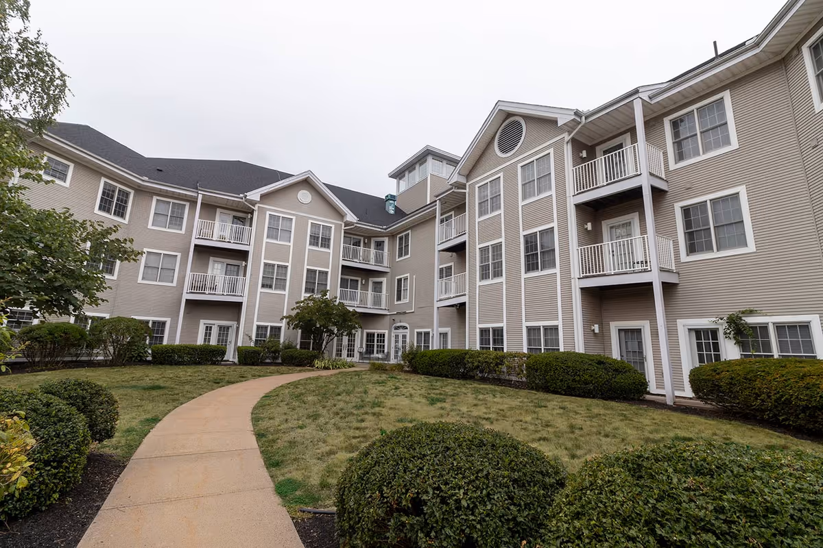 Exterior view of a multi-story beige residential building with balconies, a curved walkway, and landscaped lawn.