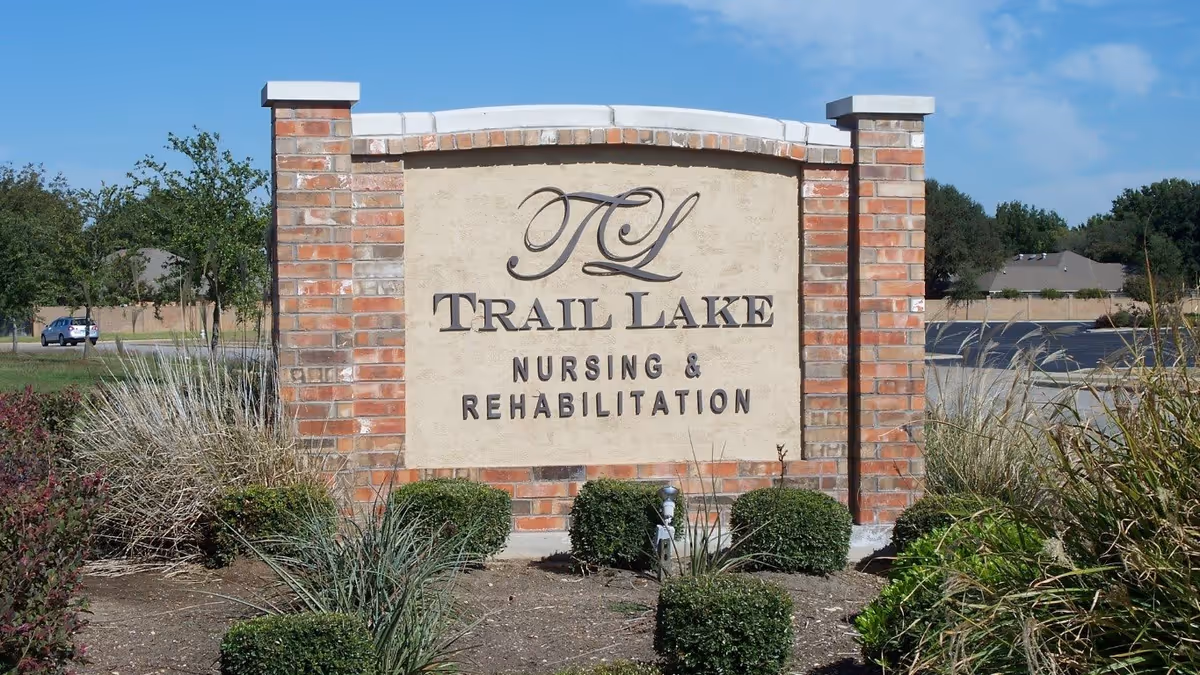 A large brick and stone sign for Trail Lake Nursing & Rehabilitation surrounded by landscaping with bushes and grass, with a parking lot and trees in the background under a clear blue sky.