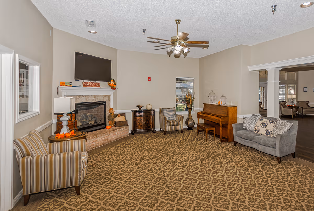 A communal sitting room with a fireplace and wall-mounted TV, piano, armchairs, and a sofa.