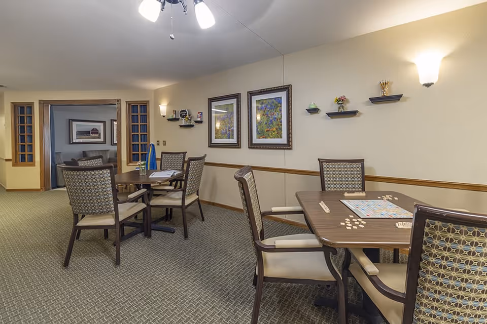 A cozy common area with two wooden tables and several cushioned chairs arranged around them. One table has a Scrabble game set up with tiles and racks. The walls are decorated with framed floral artwork and small shelves holding decorative items. The room has beige walls, patterned carpet, and soft lighting from wall sconces and a ceiling light fixture. An adjoining room with more seating is visible through a wide doorway.