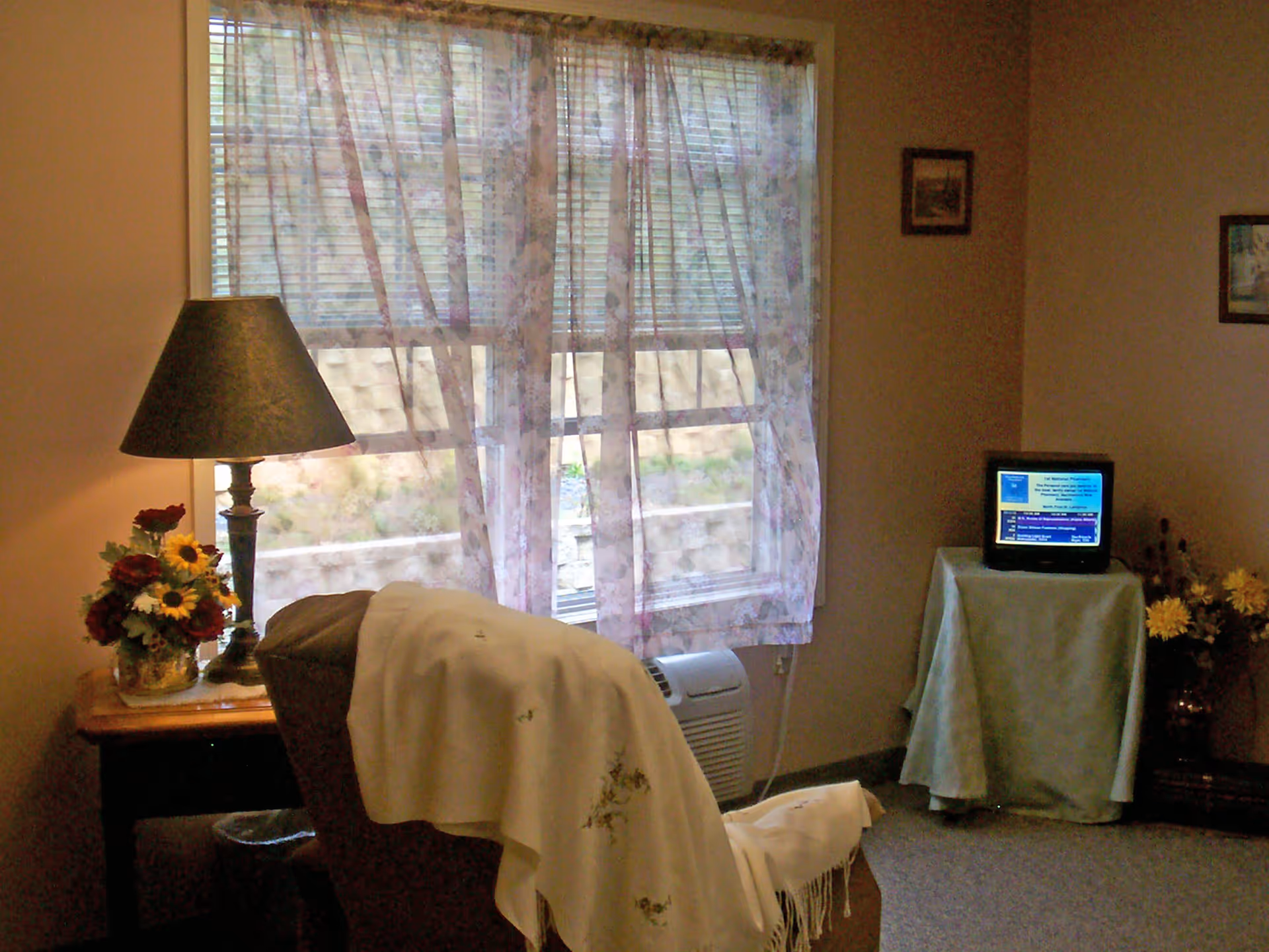A cozy living room area with a large window covered by sheer floral curtains. In front of the window is a comfortable armchair draped with a white throw blanket. To the left of the chair is a wooden side table with a lamp and a vase of flowers. On the right side of the room, a small TV sits on a table covered with a cloth, and there are framed pictures on the walls.