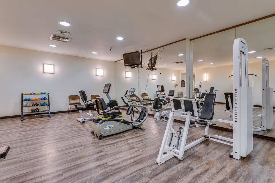 A fitness room with exercise equipment including stationary bikes, weight machines, and a rack of colorful dumbbells. The room has wood flooring, white walls with mounted lights, and a large mirror covering one wall. Two televisions are mounted on the mirrored wall.