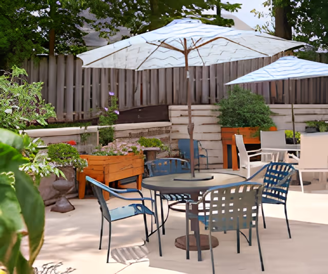 Outdoor patio with metal table and chairs under large umbrellas, planter boxes, and a wooden fence in the background.