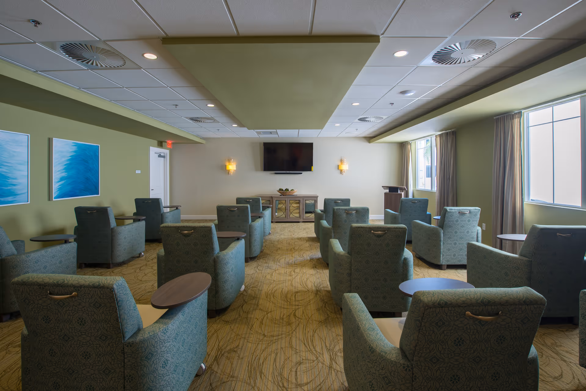 A well-lit common room with multiple green patterned armchairs arranged in rows facing a wall-mounted flat screen TV. The room has green and beige walls, carpeted floor, two large windows with curtains on the right side, and two blue abstract paintings on the left wall. There are small round tables beside some chairs and a wooden cabinet beneath the TV.