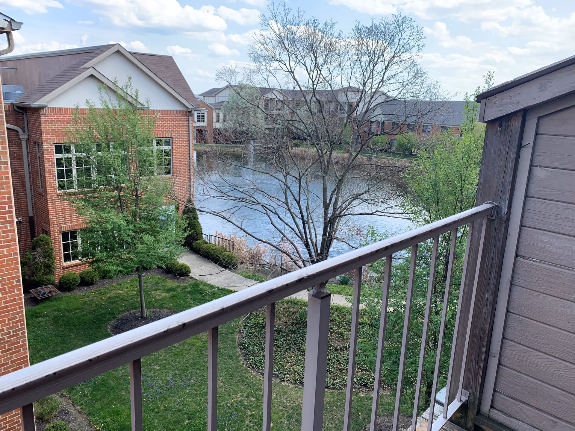 View from a balcony overlooking a landscaped garden area with green grass, trees, and shrubs. There is a brick building on the left side and a pond with calm water in the background. The sky is partly cloudy.