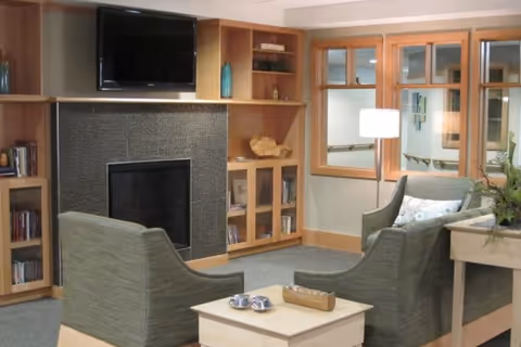 A cozy living room area in a senior living facility with two green upholstered armchairs facing a small wooden coffee table. Behind the chairs is a fireplace with a dark tiled surround and a flat-screen TV mounted above it. Wooden built-in shelves and cabinets flank the fireplace, holding books and decorative items. A window with wooden trim and a lamp is visible in the background.