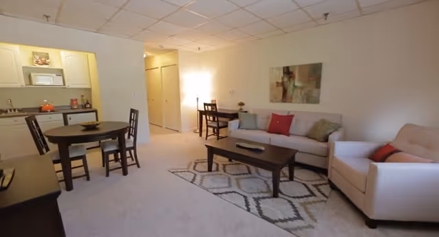 A cozy living area in a senior living facility featuring a beige sofa with colorful cushions, a matching armchair, a dark wooden coffee table on a patterned rug, a small dining table with four chairs, and a kitchenette with white cabinets and appliances in the background.