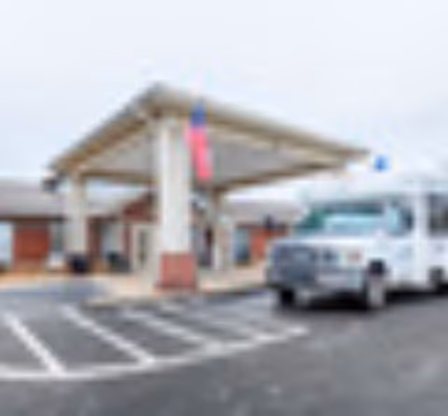 Exterior view of Wellington Manor Jackson showing the entrance with a covered drop-off area supported by columns. An American flag is hanging from the structure, and a white shuttle bus is parked nearby in the parking lot.