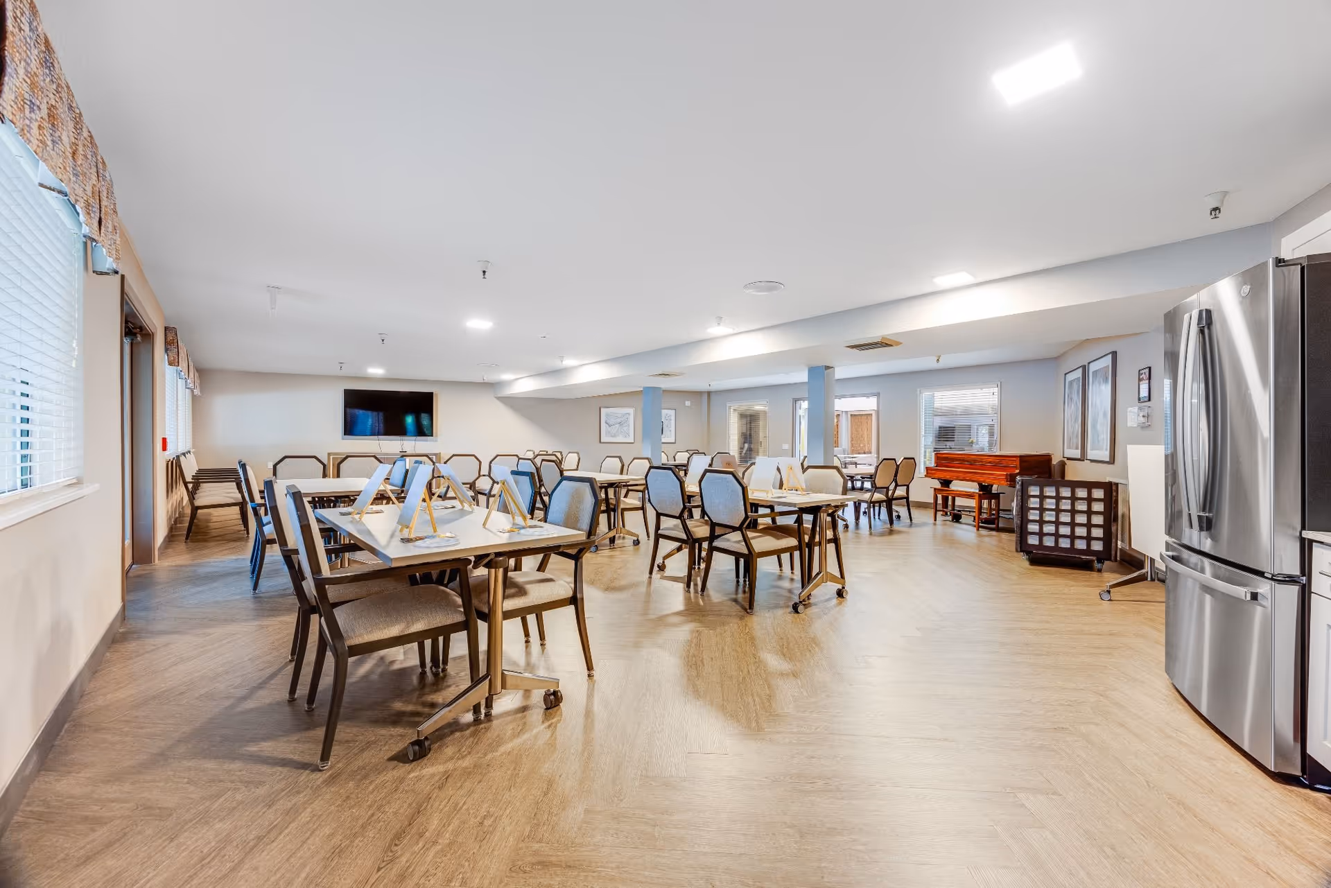 Spacious communal dining room with multiple tables and chairs, a TV on the far wall and a refrigerator on the right.