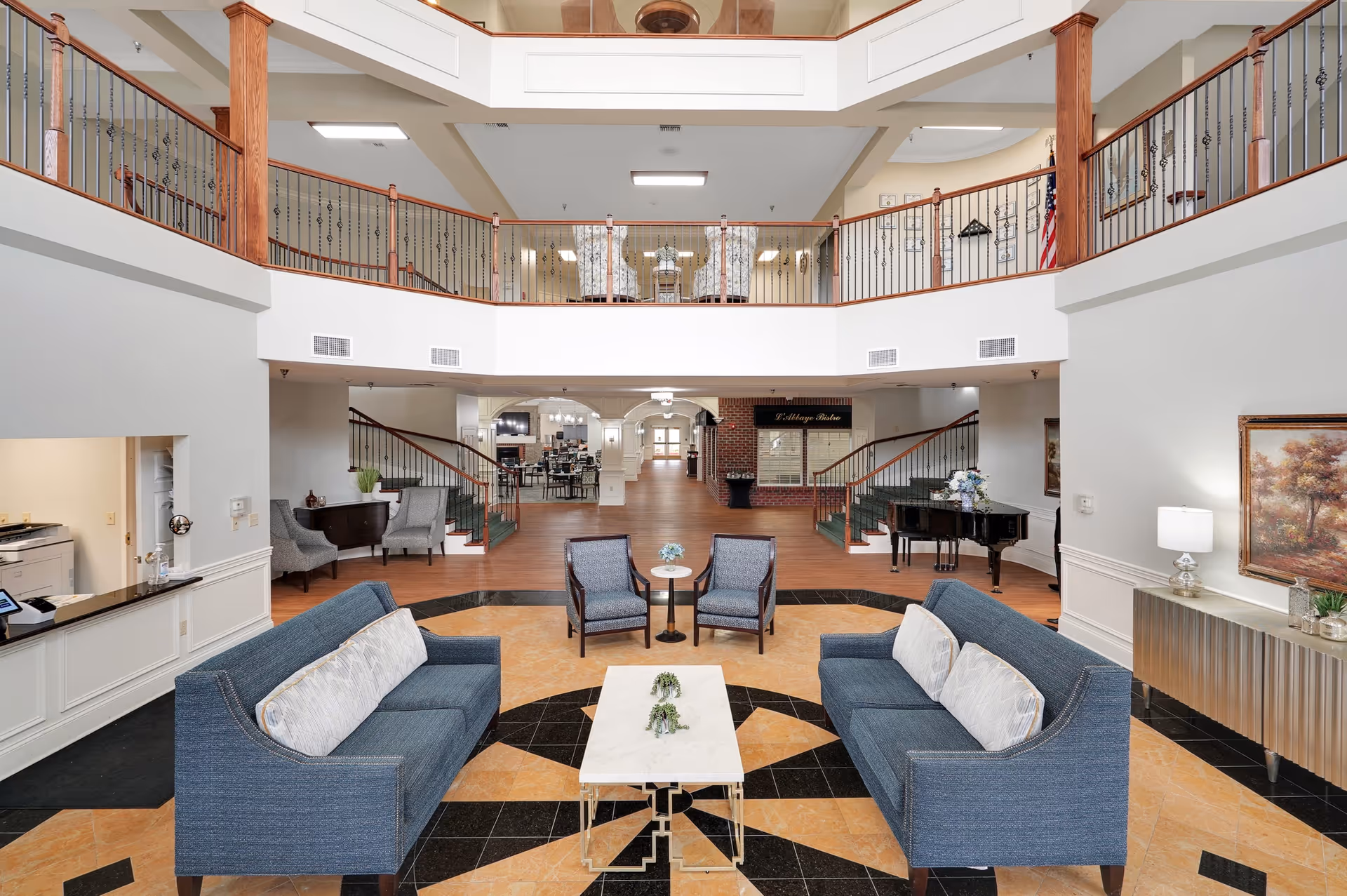 Spacious senior living facility common area with two blue sofas facing each other across a white coffee table, two armchairs, and a grand piano in the background. The room features a high ceiling with a balcony railing on the second floor, wood flooring, and neutral-colored walls with decorative artwork and lamps.
