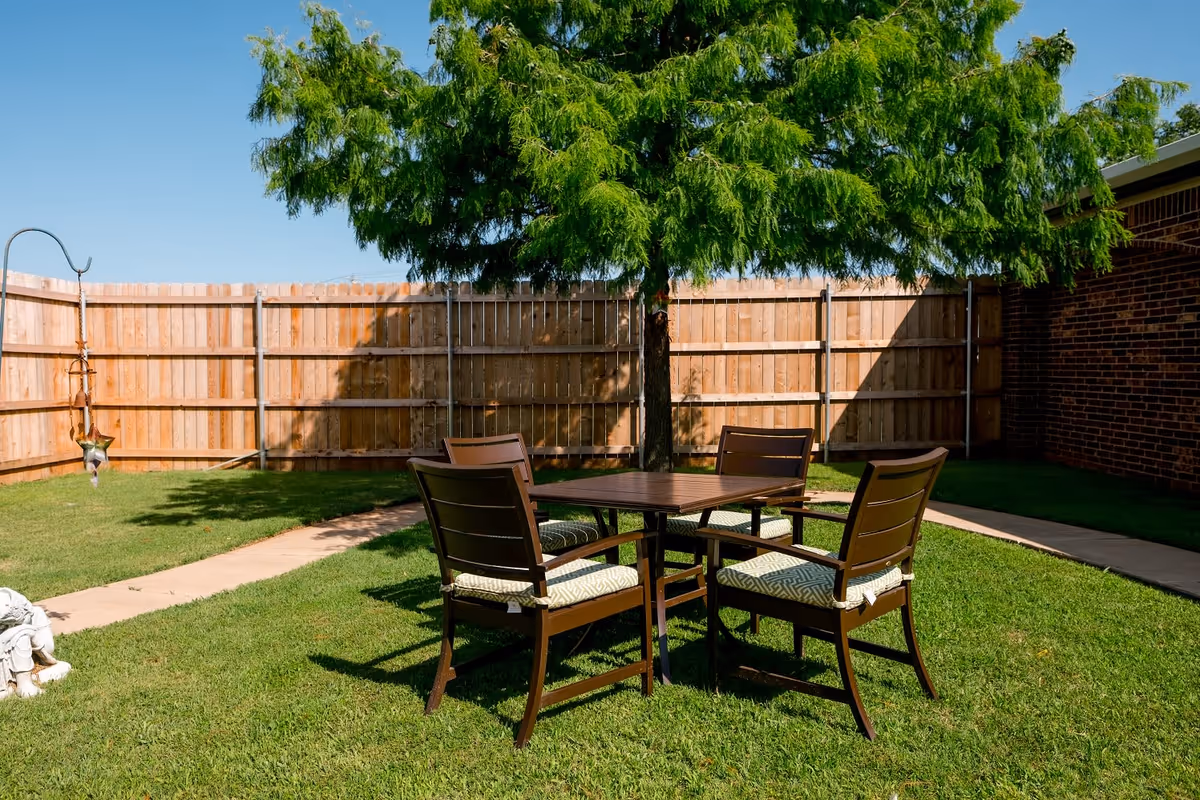 Outdoor patio area with a wooden table and four cushioned chairs on green grass under a large leafy tree, surrounded by a wooden fence and a brick building wall on the right side.