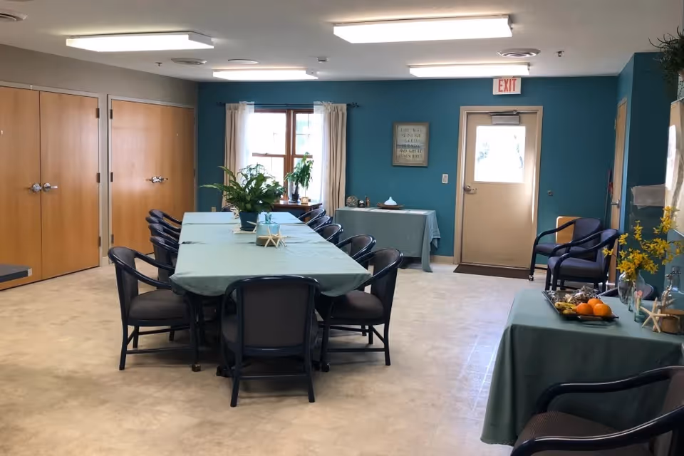 A community dining/activity room with two long teal-covered tables surrounded by chairs, potted plants, seating against teal walls, and a door at the far end.