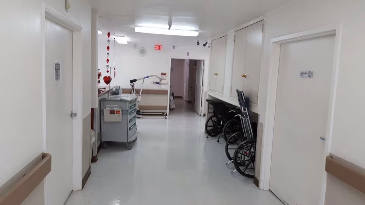 A bright clinical hallway with wheelchairs and a medical supply cart, doors on both sides and an exit sign at the far end.