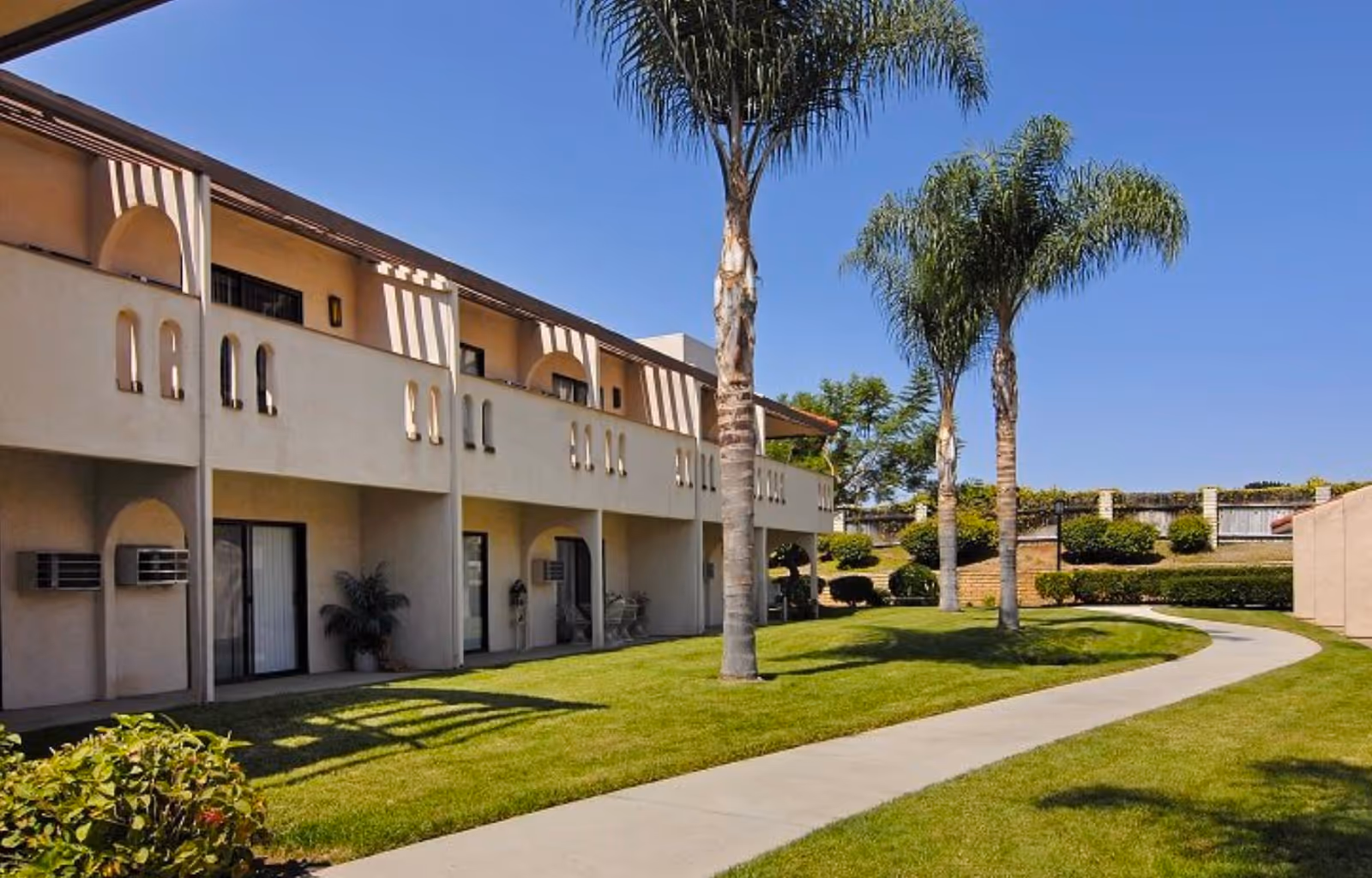 Two-story beige senior living building with balconies overlooking a curved walkway, palm trees, and a manicured lawn.