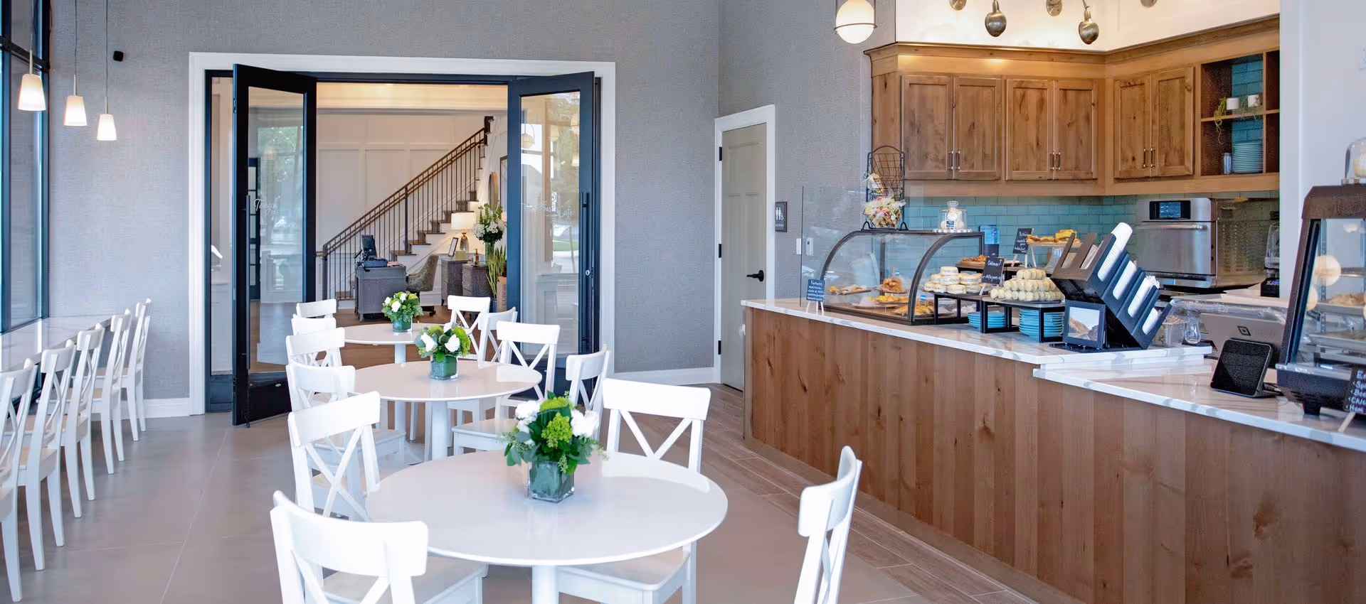 Bright and clean dining area with white round tables and chairs, each table decorated with a small green and white floral arrangement. To the right, there is a wooden counter with a glass display case containing pastries and other baked goods. Behind the counter are wooden cabinets and kitchen appliances. Large glass doors open to a hallway with stairs and seating visible in the background.