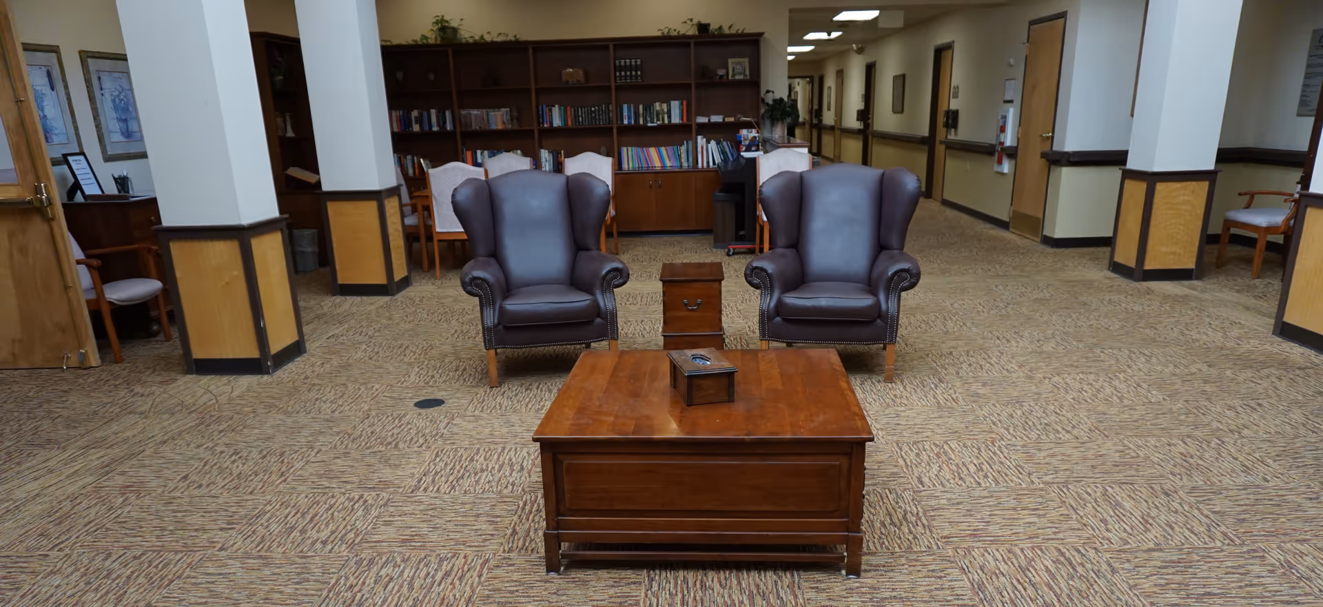 Carpeted communal lounge with two leather wingback chairs facing a wooden coffee table and bookshelves in the background.