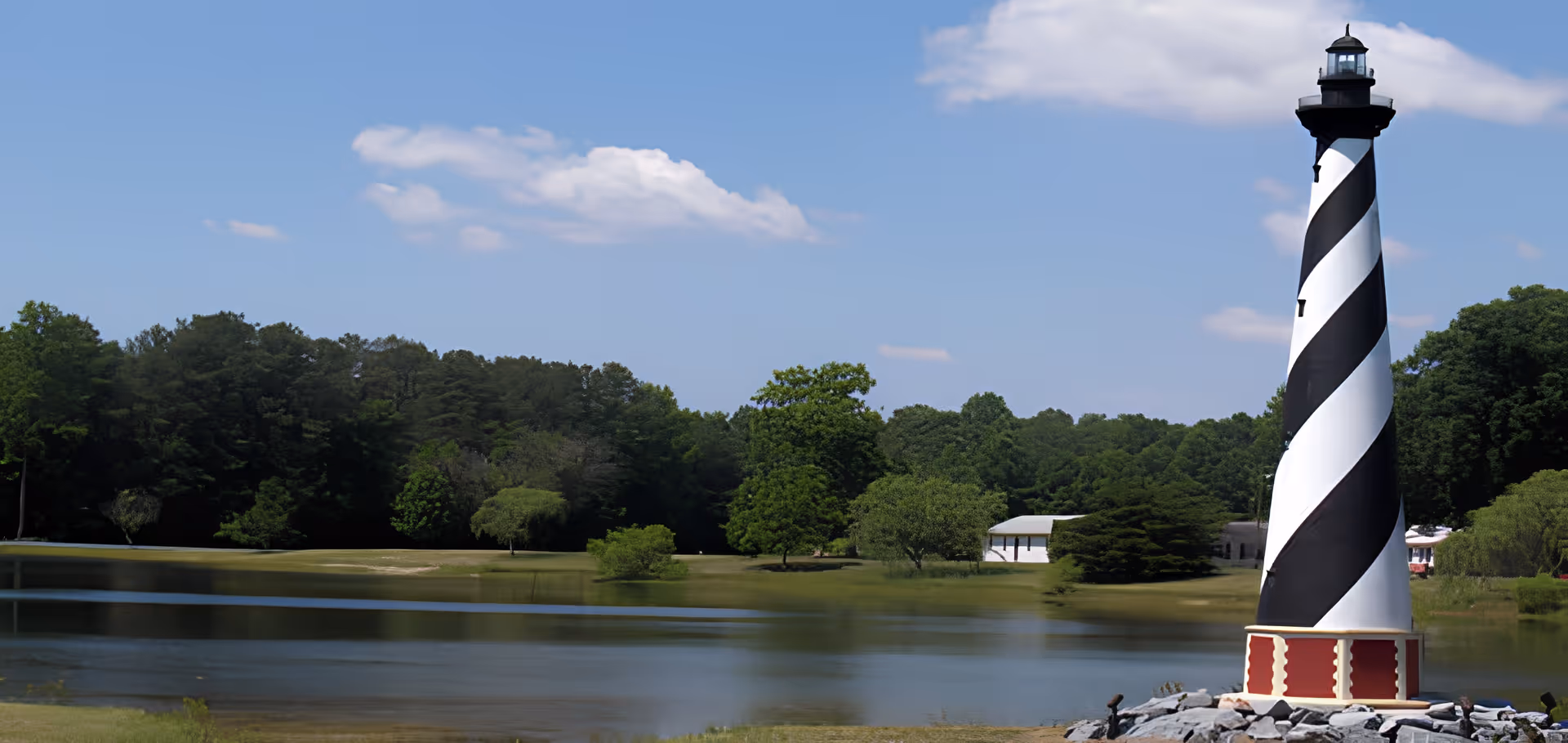 A black and white striped lighthouse stands on a rocky base near a calm body of water with green trees and a small white building in the background under a blue sky with some clouds.