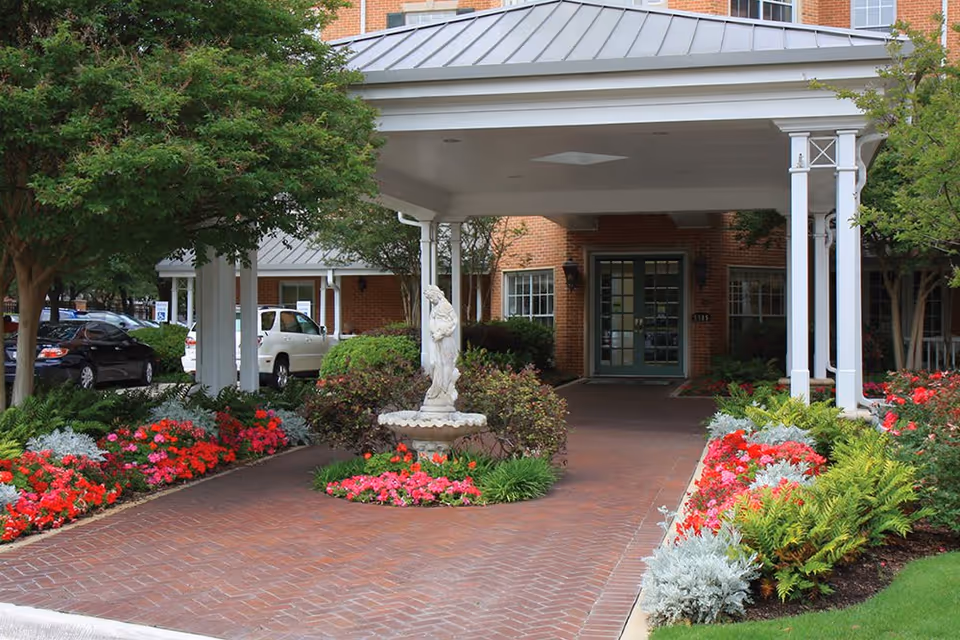 Entrance to Caruth Haven Court facility with a covered driveway, a white statue fountain in the center surrounded by colorful flower beds and greenery, and a brick building with green double doors in the background.