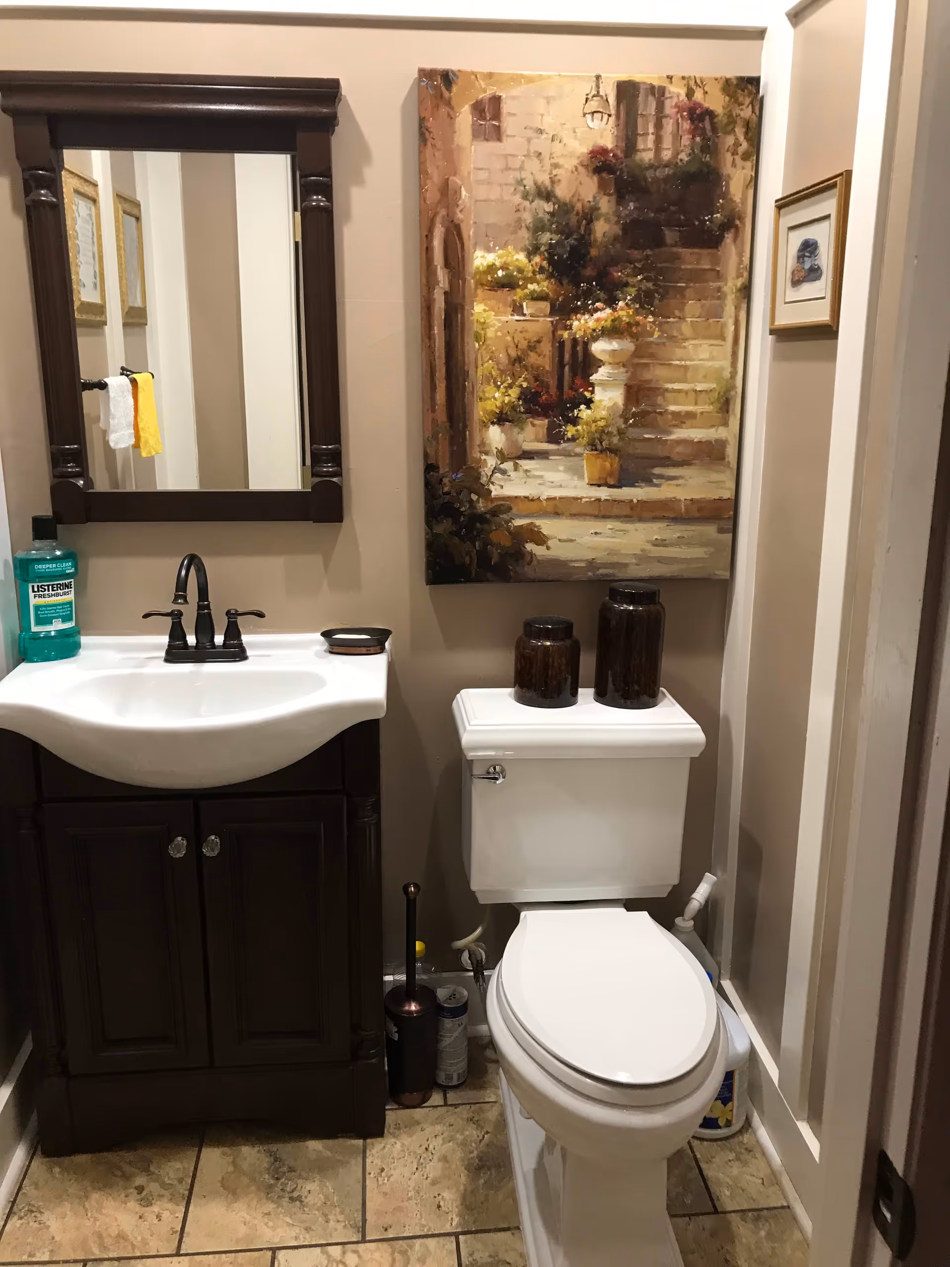 A small bathroom with a white toilet and a dark wood vanity with a white sink. Above the sink is a dark wood framed mirror. On the toilet tank are two dark decorative jars. A painting of a stone staircase with flowers hangs on the wall above the toilet. A bottle of Listerine mouthwash is on the sink counter, and a toilet brush and cleaning supplies are on the floor beside the toilet.