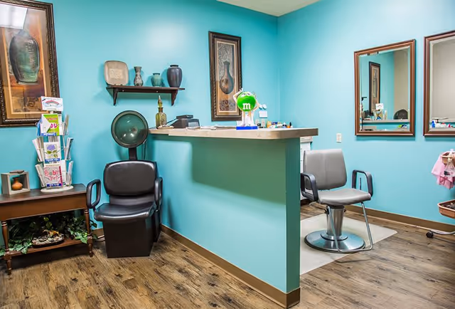 Interior of a salon area with turquoise walls, featuring a black salon chair with a hair dryer hood, a gray salon chair in front of two mirrors, a small counter with decorative items, and a wooden side table with brochures and plants underneath.