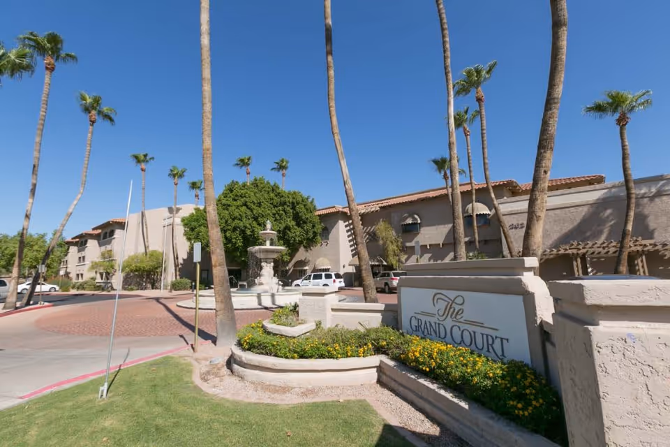 Exterior view of The Grand Court senior living facility with palm trees, a fountain, parked cars, and a clear blue sky.