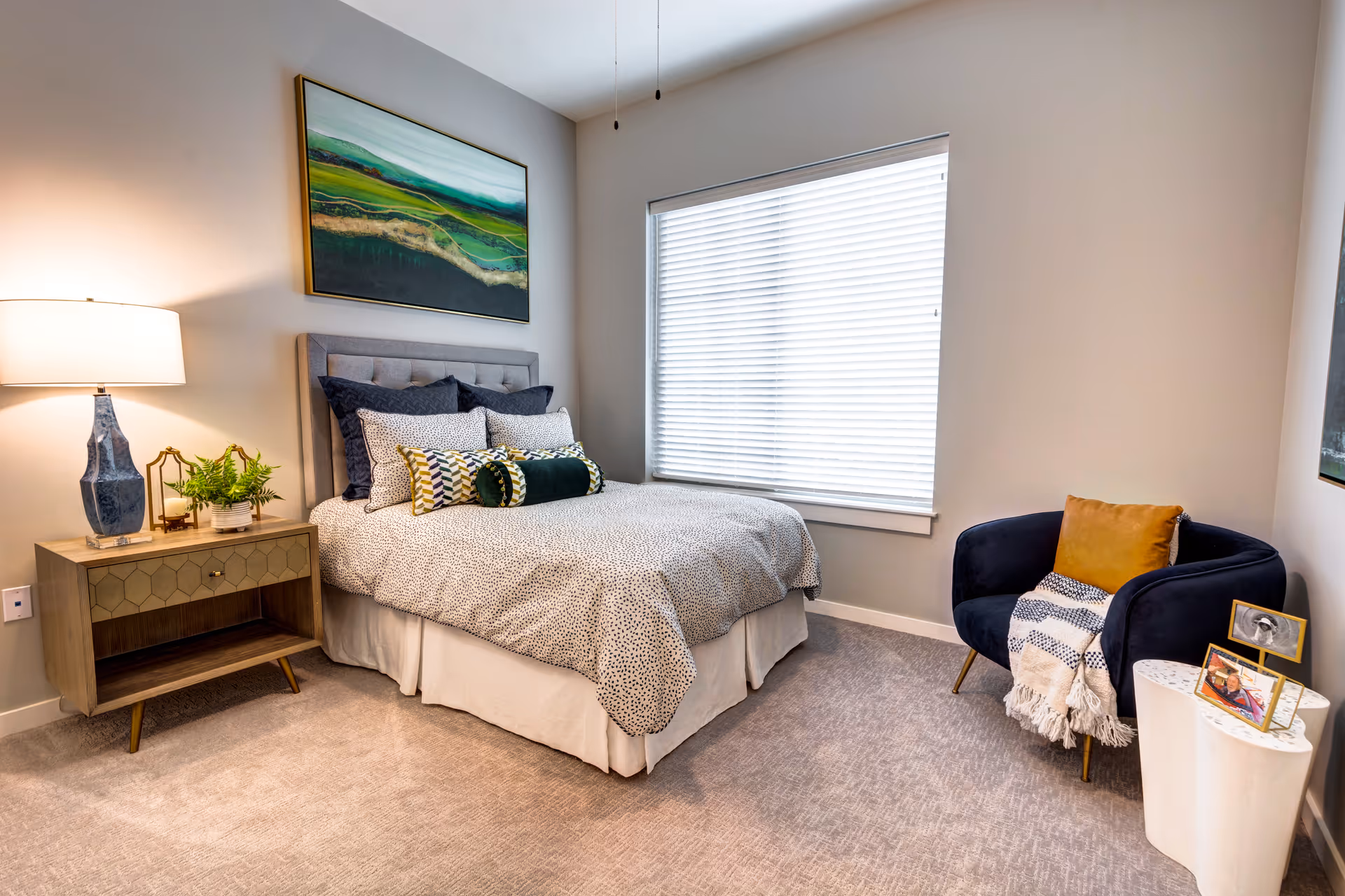 A cozy bedroom with a bed featuring multiple pillows and a patterned bedspread. To the left of the bed is a wooden nightstand with a blue lamp, a small plant, and decorative items. Above the bed hangs a landscape painting with green and blue tones. To the right, there is a navy blue armchair with a mustard yellow pillow and a striped throw blanket. Next to the chair is a small white side table with two framed photos. The room has a large window with white blinds and light-colored carpet flooring.