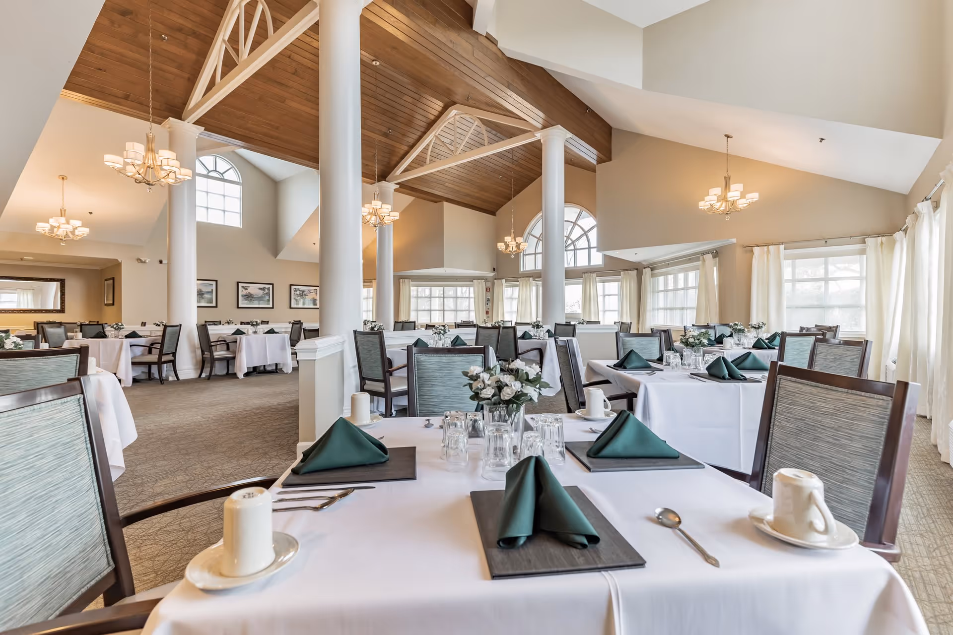 A spacious dining room with high vaulted ceilings and wooden beams. The room features multiple tables covered with white tablecloths, each set with green folded napkins, glassware, and white coffee cups. Large windows with sheer curtains allow natural light to fill the room. Elegant chandeliers hang from the ceiling, and white columns add architectural interest.
