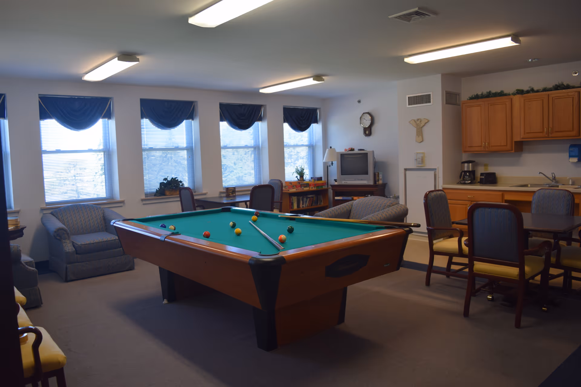 Interior recreation room with pool table, sofas, a kitchenette, and windows with blue valances.