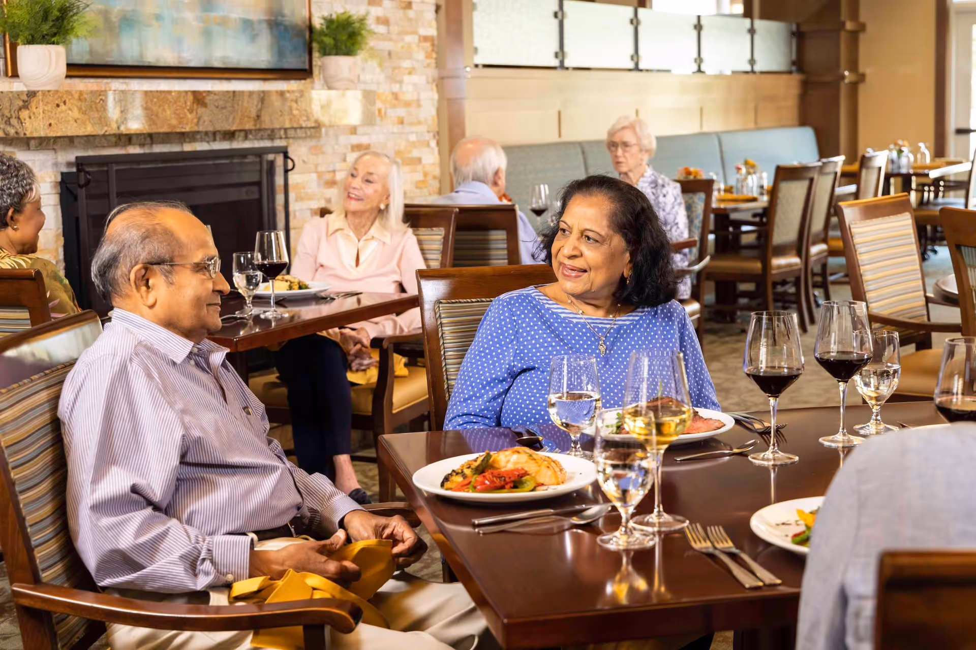 Older adults sit at a communal dining table in a bright, restaurant-style dining room enjoying plated meals and wine.