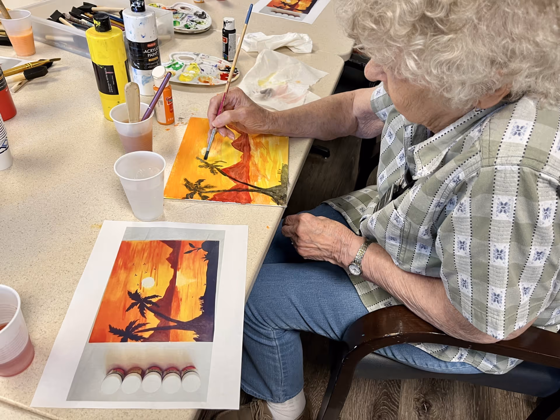An elderly woman with curly white hair is seated at a table, painting a sunset scene with palm trees on a canvas. Various painting supplies, including bottles of acrylic paint, paintbrushes, and a reference image of the sunset scene, are spread out on the table.
