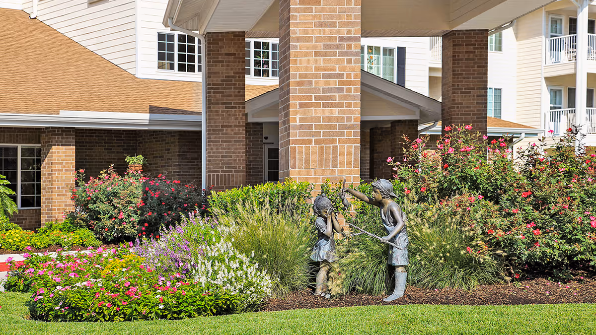 A landscaped garden area in front of a brick building with a covered entrance supported by large brick columns. The garden features colorful flowers, green shrubs, and a bronze statue of two children playing. The building has multiple windows and a tan roof.