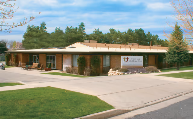 Single-story brick building with a flat roof surrounded by green grass, trees, and a paved driveway. There is outdoor seating with chairs and tables near the entrance. A sign in front of the building reads 'Willow Glen Health and Rehab.'